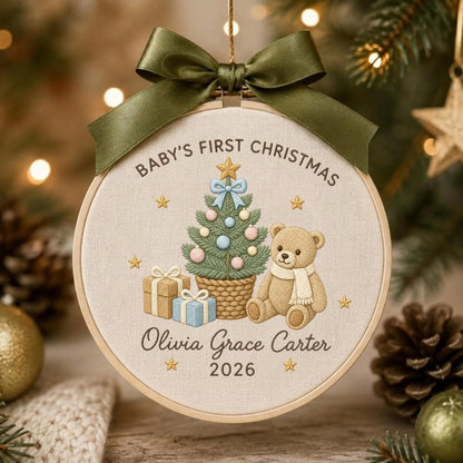 May include: A round embroidery hoop ornament featuring a beige fabric background with a green ribbon bow at the top. The design includes a Christmas tree, a teddy bear, gifts, and the text Baby's First Christmas, Olivia Grace Carter, and 2026.
