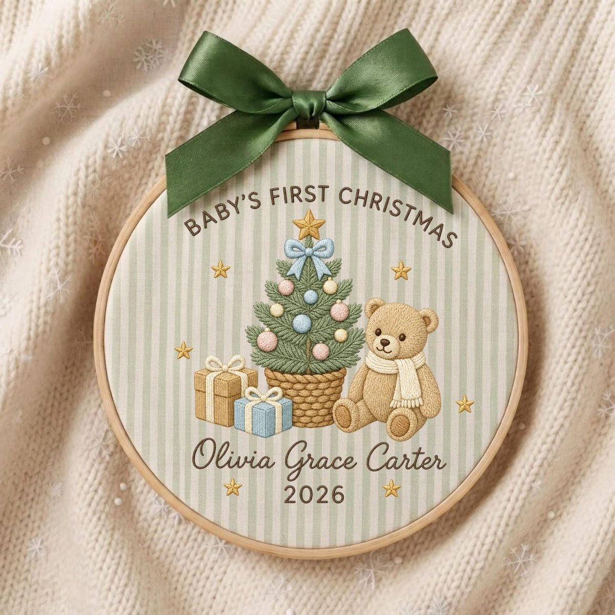 May include: A round wooden embroidery hoop featuring a light green and white striped fabric. The design includes a decorated Christmas tree, a teddy bear, and gifts. Text reads: Baby's First Christmas, Olivia Grace Carter, 2026. A dark green ribbon bow is attached to the top.