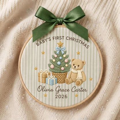 May include: A round wooden embroidery hoop featuring a light green and white striped fabric. The design includes a decorated Christmas tree, a teddy bear, and gifts. Text reads: Baby's First Christmas, Olivia Grace Carter, 2026. A dark green ribbon bow is attached to the top.