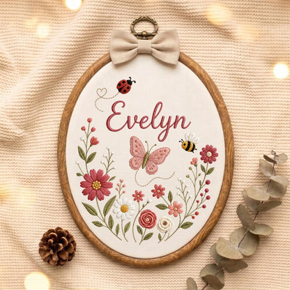 May include: An oval wooden embroidery hoop containing white fabric with the embroidered name Evelyn in dark pink script. The design features a pink butterfly, a red ladybug, a yellow and black bee, and various pink, white, and green flowers.