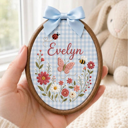 May include: An oval wooden embroidery hoop featuring a light blue and white gingham fabric background. The fabric is embroidered with the name Evelyn in red script, a pink butterfly, a ladybug, a bee, and various colorful floral stems.