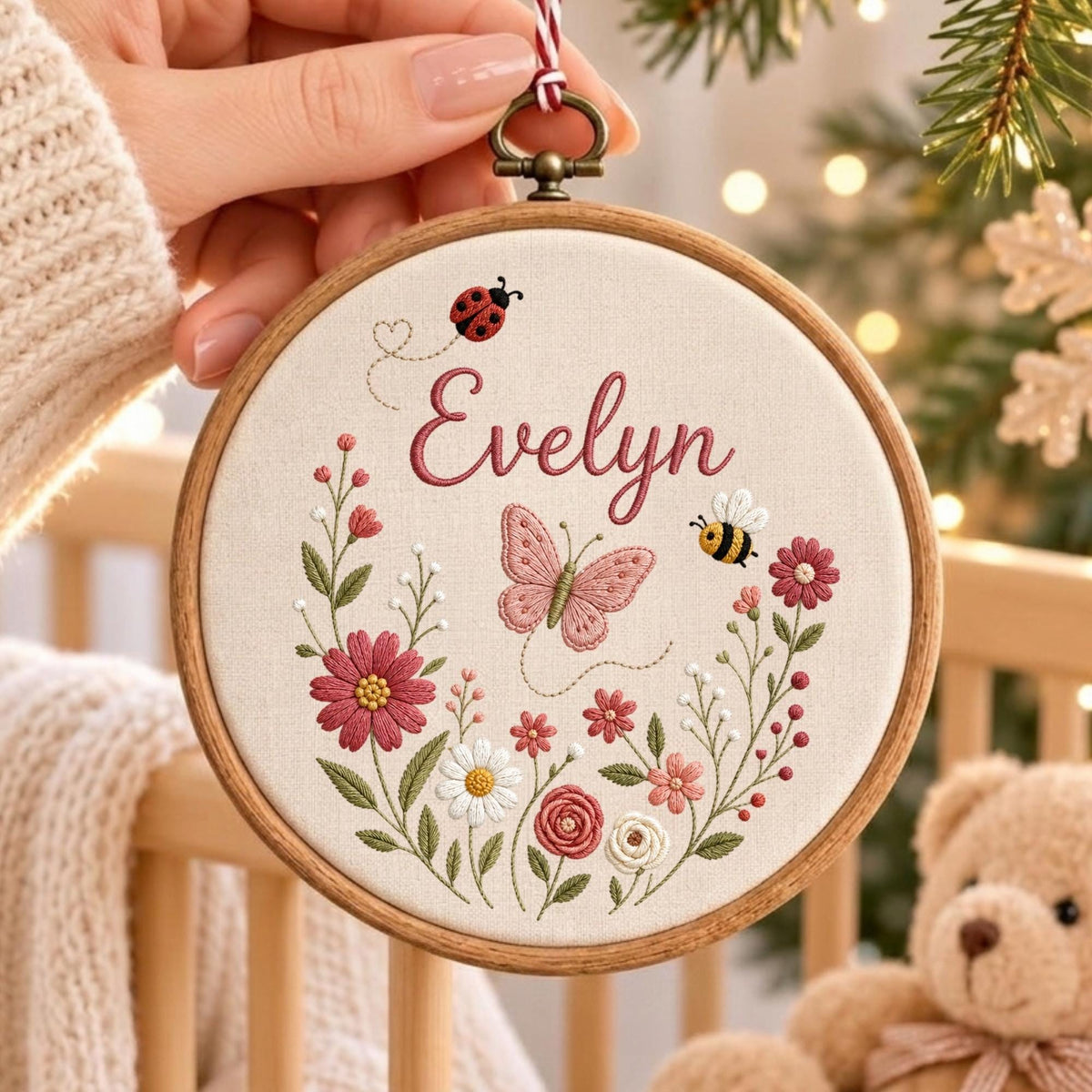 May include: A circular wooden embroidery hoop containing cream-colored fabric with the name Evelyn stitched in pink. The design features a pink butterfly, a ladybug, a bee, and a floral arrangement of pink and white flowers with green leaves.
