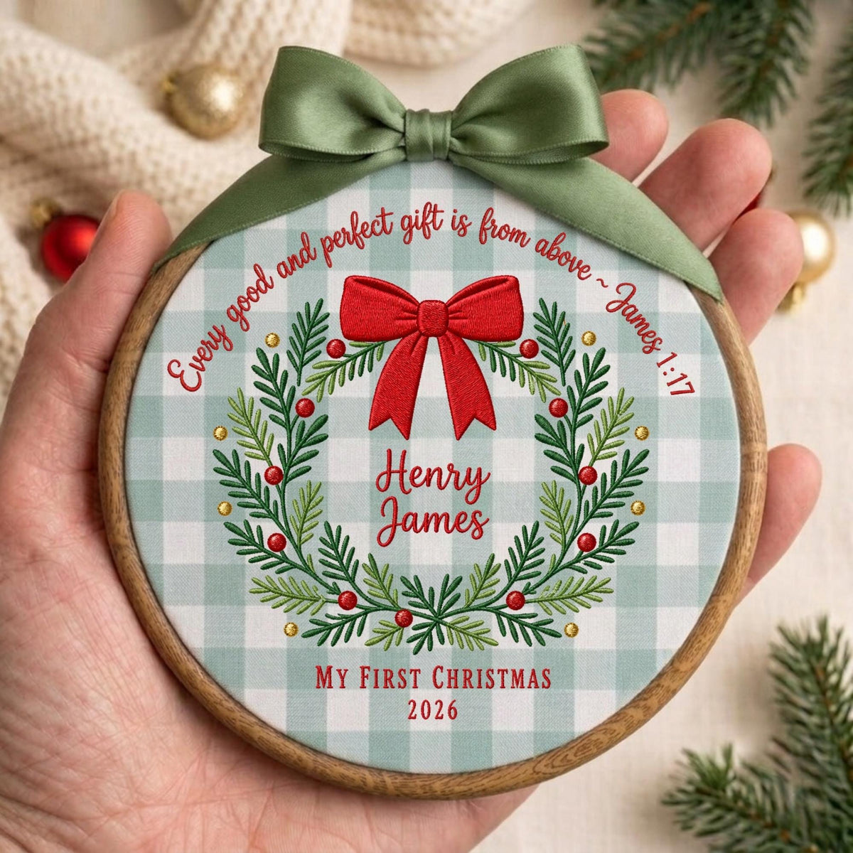 May include: A round wooden embroidery hoop ornament featuring a light green and white gingham fabric background. It displays a green wreath with red berries, a large red bow, and the text "Every good and perfect gift is from above ~ James 1:17", "Henry James", "My First Christmas", and "2026". An olive green satin ribbon is tied at the top.