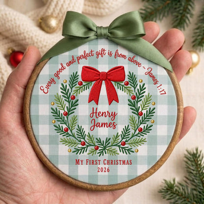 May include: A round wooden embroidery hoop ornament featuring a light green and white gingham fabric background. It displays a green wreath with red berries, a large red bow, and the text "Every good and perfect gift is from above ~ James 1:17", "Henry James", "My First Christmas", and "2026". An olive green satin ribbon is tied at the top.