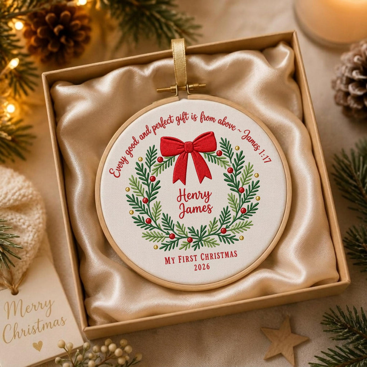 May include: A round wooden embroidery hoop ornament featuring white fabric with a green embroidered wreath, a red bow, and red text reading Every good and perfect gift is from above ~ James 1:17, Henry James, and MY FIRST CHRISTMAS 2026.
