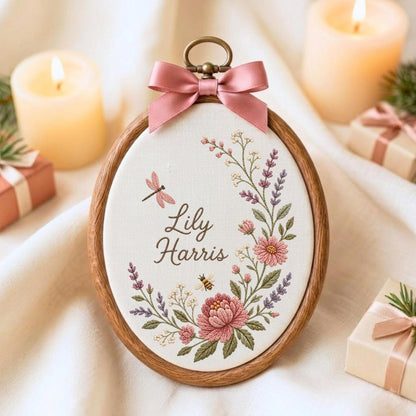 May include: An oval embroidery hoop featuring a white fabric background with the embroidered name Lily Harris, surrounded by a floral wreath of pink flowers, lavender sprigs, a dragonfly, and a bee, topped with a dusty pink satin ribbon bow.