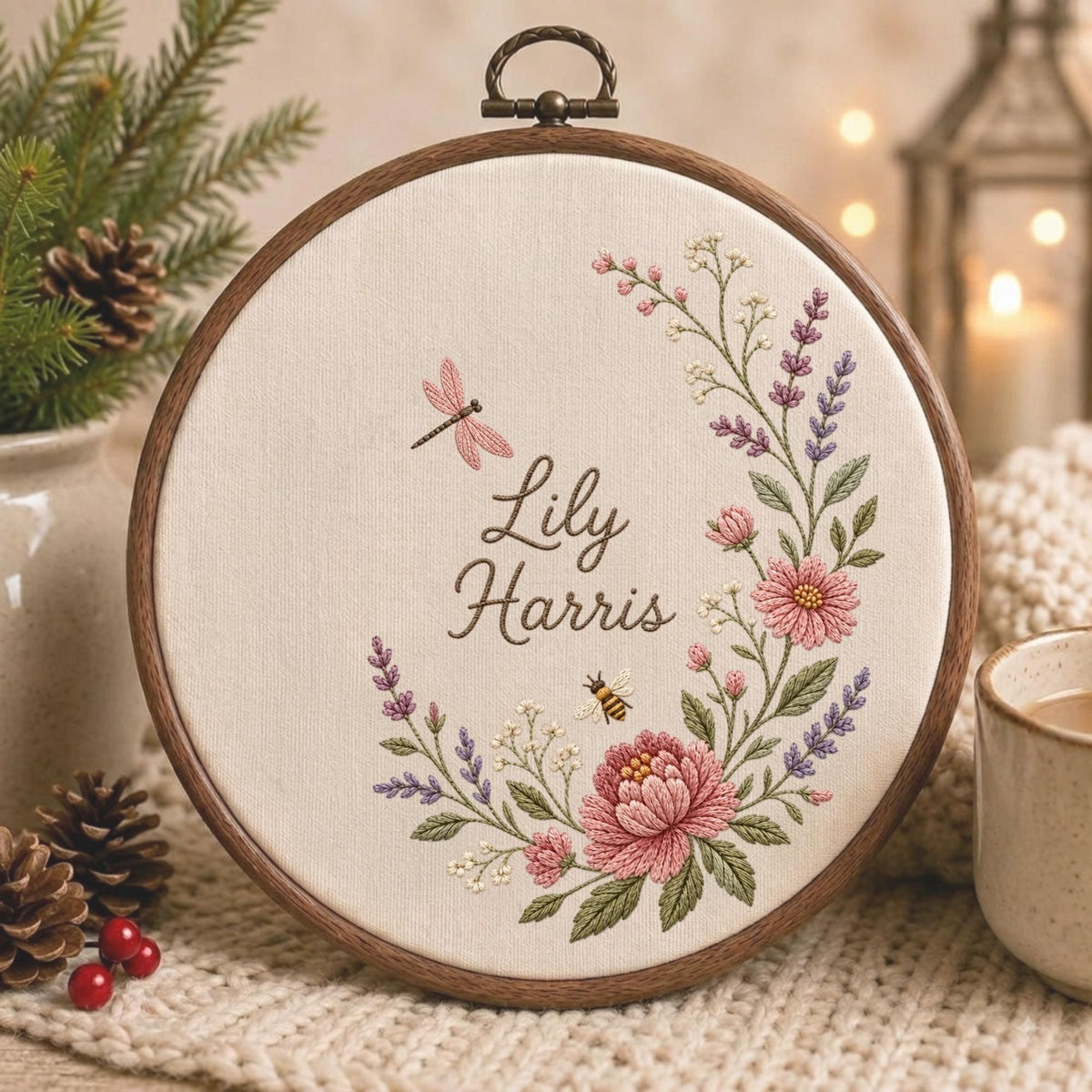 May include: A circular wooden embroidery hoop containing off-white fabric with the embroidered name Lily Harris in brown script. The design features a floral wreath of pink flowers, purple lavender, green leaves, a pink dragonfly, and a small bee.