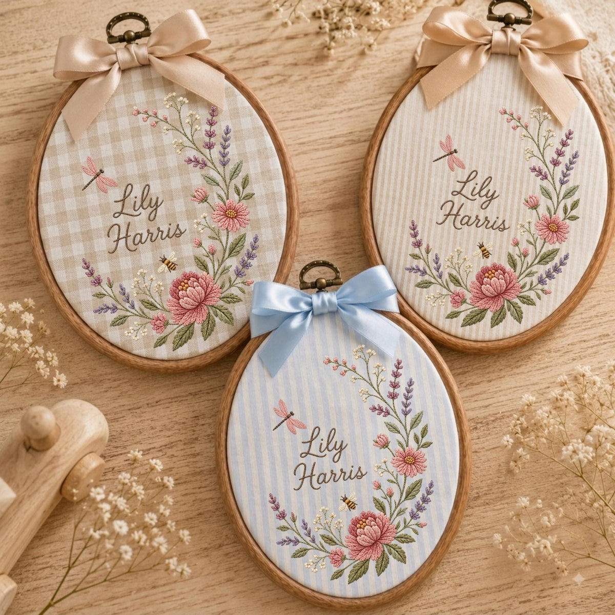 May include: Three oval wooden embroidery hoops featuring fabric backgrounds with gingham and striped patterns. Each hoop displays embroidered floral wreaths, a dragonfly, a bee, and the name Lily Harris, topped with a decorative satin bow.