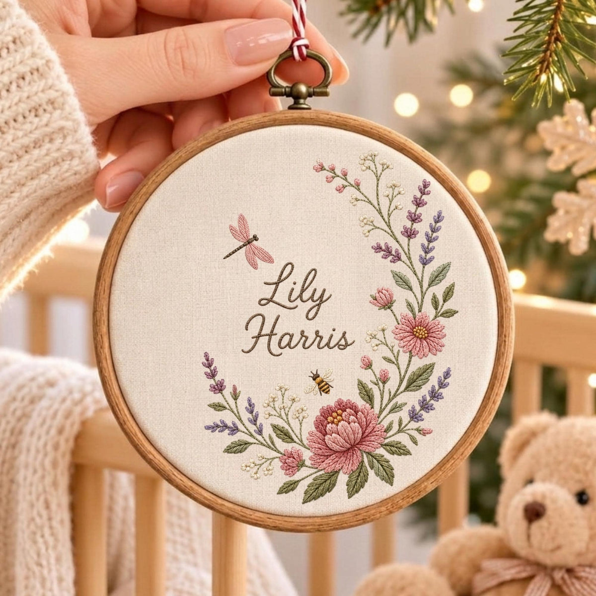May include: A circular wooden embroidery hoop containing white fabric with the embroidered name Lily Harris. The design features a floral wreath with pink flowers, purple lavender, green leaves, a small dragonfly, and a bee in the center.