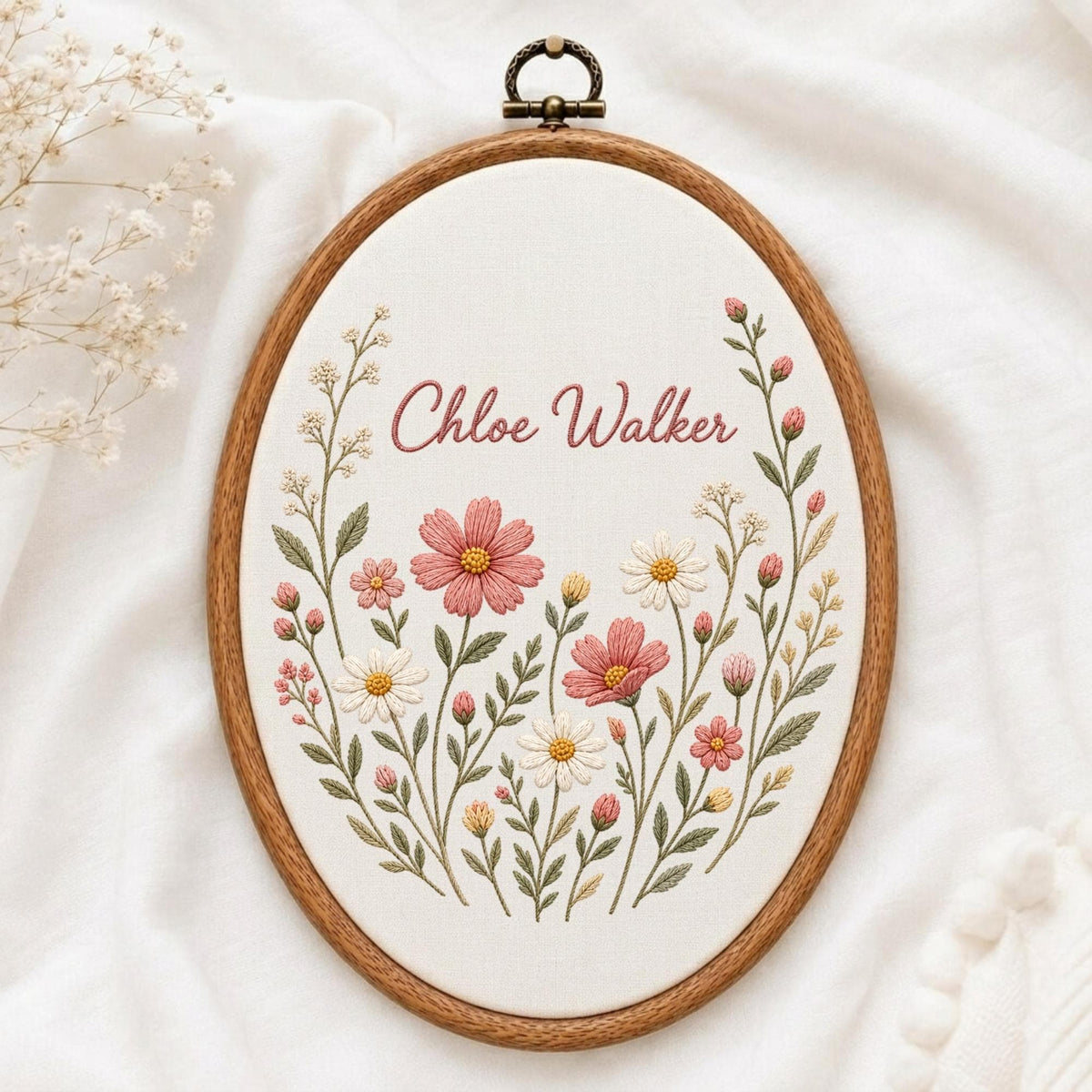 May include: An oval embroidery hoop featuring a wooden frame and a metal hanging loop at the top. The white fabric center displays hand-stitched floral motifs in shades of pink, white, and green, with the name Chloe Walker embroidered in pink.