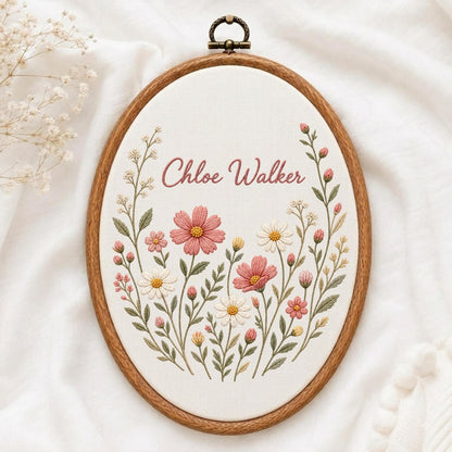 May include: An oval embroidery hoop featuring a wooden frame and a metal hanging loop at the top. The white fabric center displays hand-stitched floral motifs in shades of pink, white, and green, with the name Chloe Walker embroidered in pink.