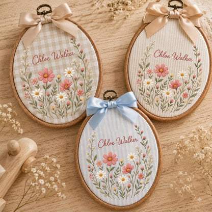 May include: Three oval wooden embroidery hoops featuring fabric backgrounds with the name Chloe Walker embroidered in cursive. Each hoop is topped with a satin ribbon bow and displays a floral arrangement of pink and white flowers with green stems.