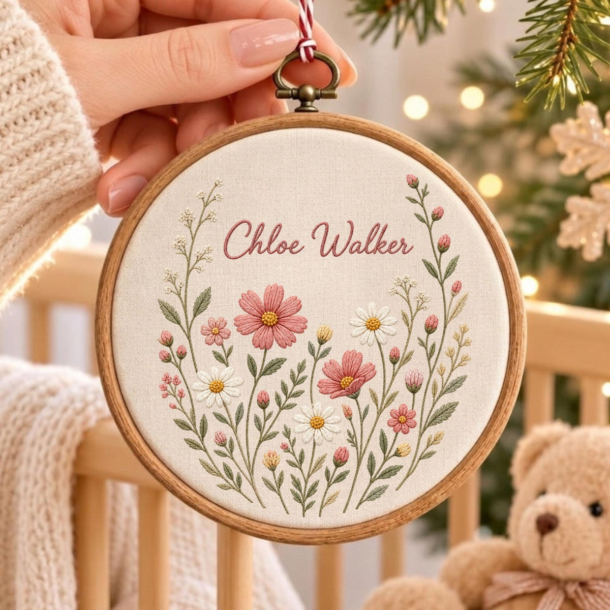 May include: A circular wooden embroidery hoop containing off-white fabric with the embroidered name Chloe Walker in pink script. The design features a floral arrangement of pink and white flowers with green stems and leaves, suspended by a red and white string.