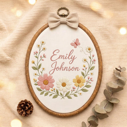 May include: An oval wooden embroidery hoop containing white fabric with the embroidered name Emily Johnson in pink script. The design features a floral wreath with pink, white, and yellow flowers, green leaves, and a small pink butterfly, topped with a beige fabric bow.