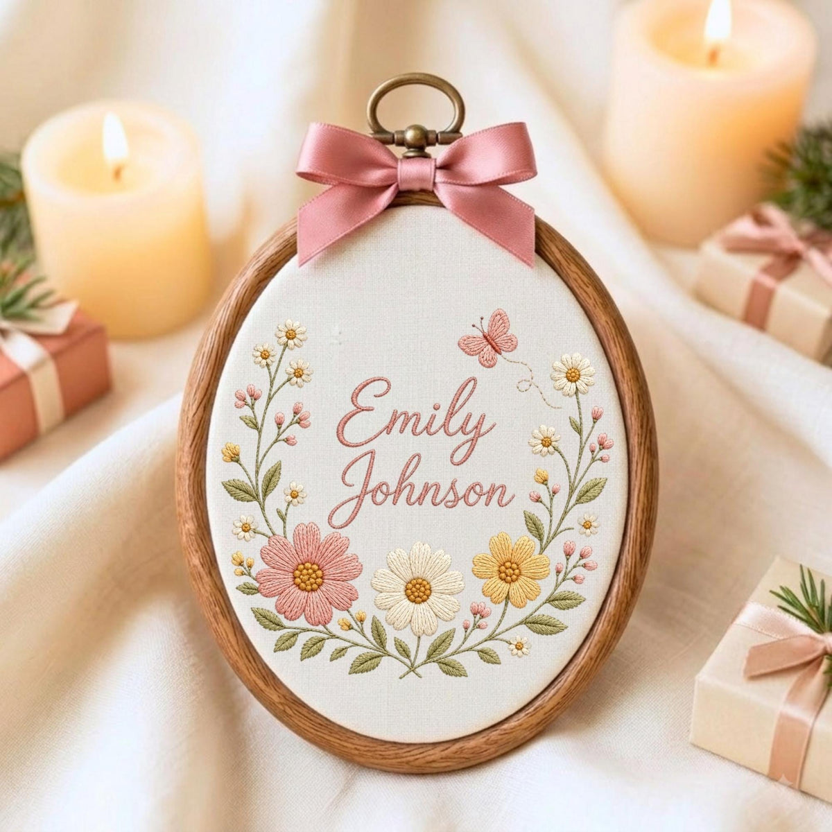 May include: An oval wooden embroidery hoop containing white fabric with the embroidered name Emily Johnson in pink script. The design features a floral wreath with pink, white, and yellow flowers, green leaves, and a small pink butterfly, topped with a pink satin bow.
