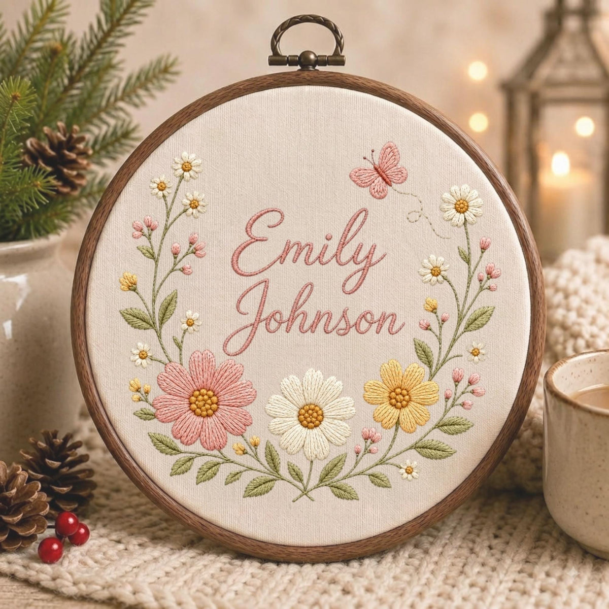 May include: A circular wooden embroidery hoop containing cream-colored fabric with the embroidered name Emily Johnson in pink script. The text is surrounded by a floral wreath featuring pink, white, and yellow flowers, green leaves, and a small pink butterfly.
