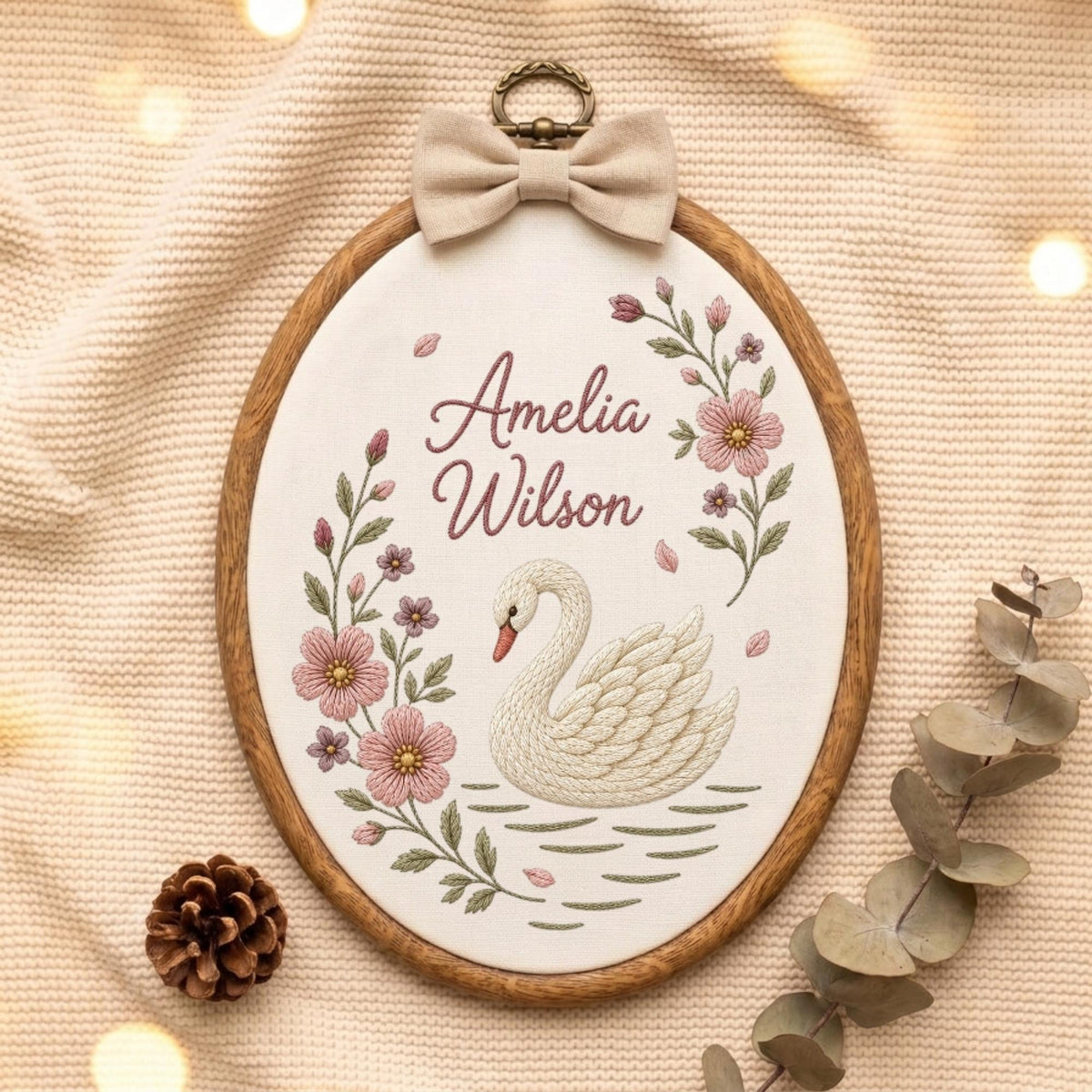 May include: An oval wooden embroidery hoop containing white fabric with the embroidered name Amelia Wilson, a white swan, and pink floral vines. A beige fabric bow is attached to the top metal hanging loop of the wooden frame.