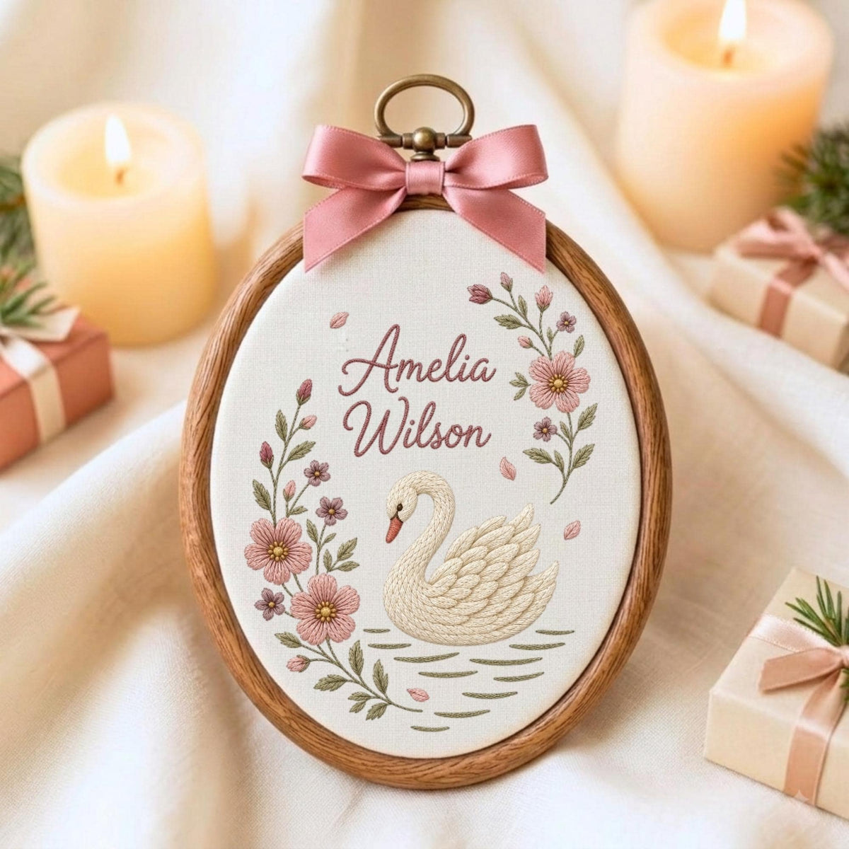 May include: An oval wooden embroidery hoop containing white fabric with a stitched white swan, pink floral vines, and the text Amelia Wilson. A dusty pink satin ribbon bow is attached to the top metal hanging loop of the embroidery frame.