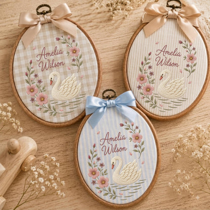 May include: Three oval wooden embroidery hoops featuring fabric backgrounds with swan and floral embroidery. Each hoop displays the name Amelia Wilson, a white swan, and pink flowers. The hoops are topped with satin bows in beige, light blue, or tan.