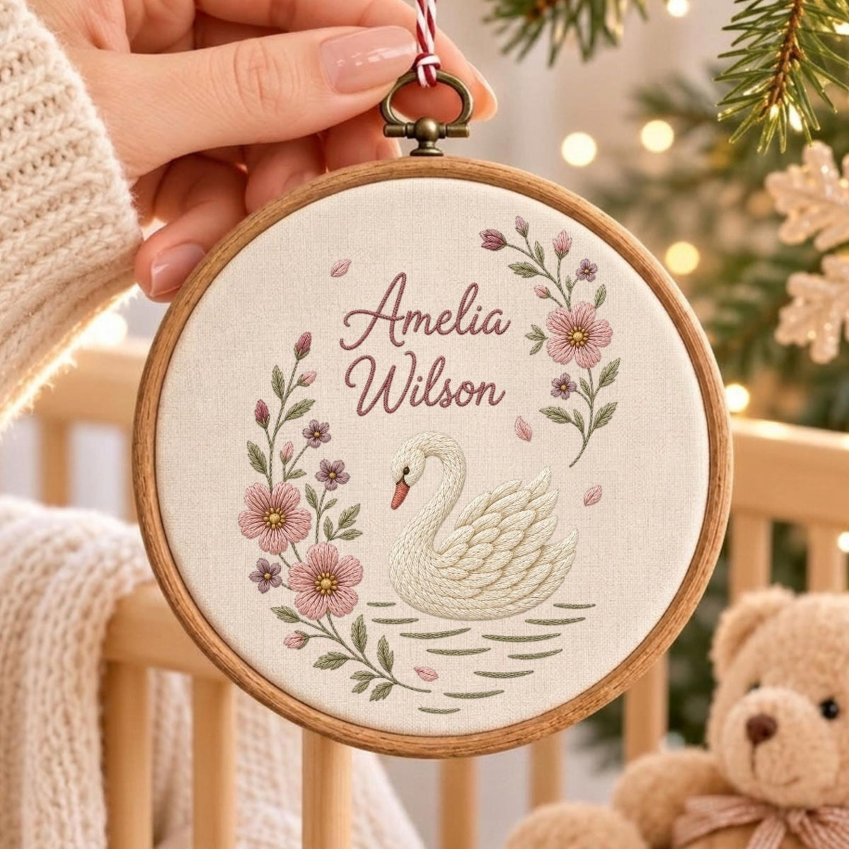 May include: A circular wooden embroidery hoop containing a cream-colored fabric panel. The fabric features an embroidered white swan, pink floral vines, and the text Amelia Wilson in a script font. A red and white string is attached to the top.