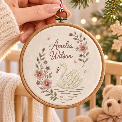 May include: A circular wooden embroidery hoop containing a cream-colored fabric panel. The fabric features an embroidered white swan, pink floral vines, and the text Amelia Wilson in a script font. A red and white string is attached to the top.