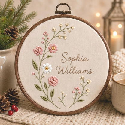 May include: A circular wooden embroidery hoop containing cream-colored fabric with hand-stitched floral embroidery in shades of pink, white, and green. The text "Sophia Williams" is embroidered in the center in a brown cursive script font.