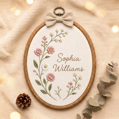May include: An oval embroidery hoop featuring a cream-colored fabric background with hand-stitched floral motifs in shades of pink, white, and green. The name Sophia Williams is embroidered in brown script. A beige fabric bow sits at the top.