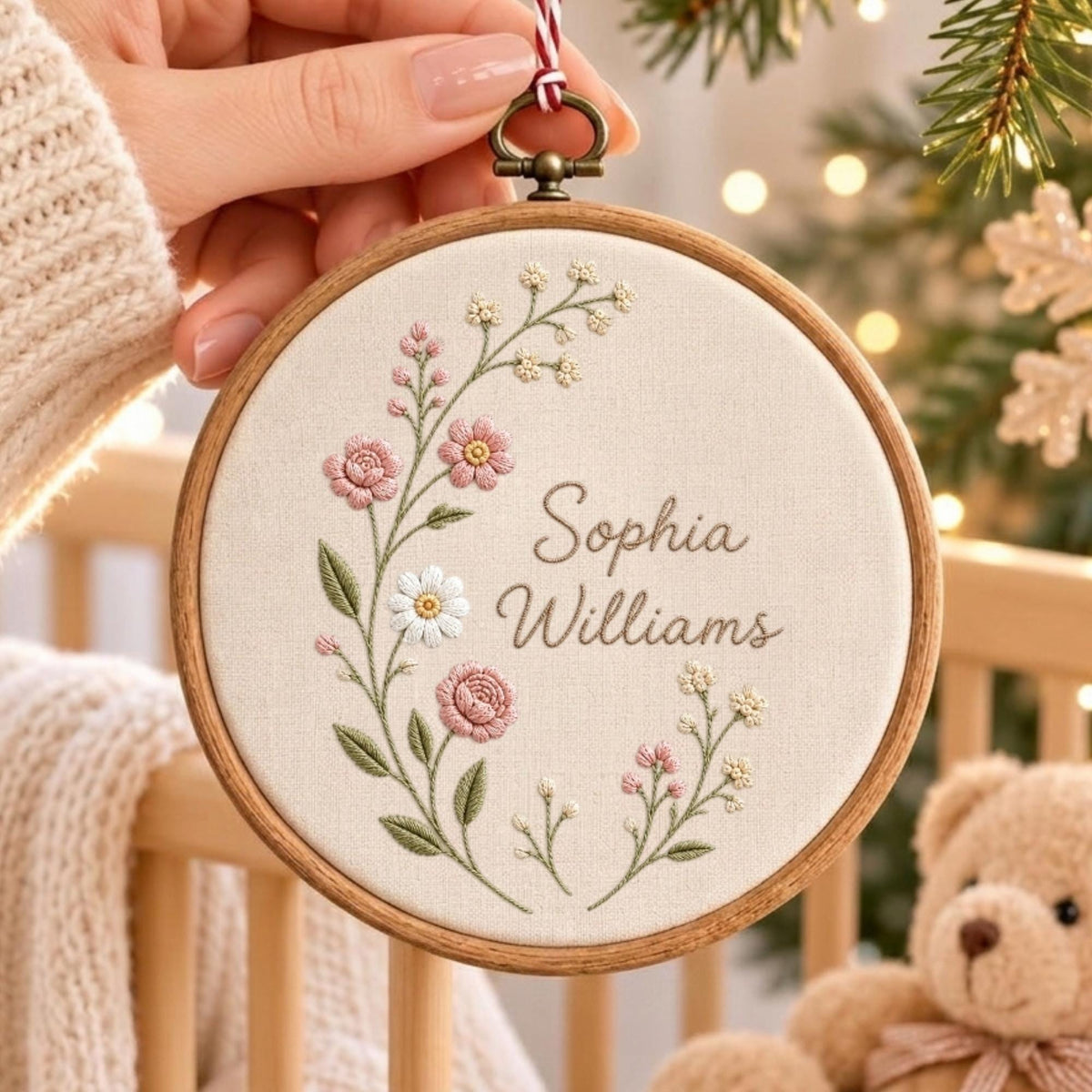 May include: A circular wooden embroidery hoop containing off-white fabric with a floral wreath design in pink, white, and green thread. The name Sophia Williams is embroidered in brown script in the center. A red and white string hangs from the top.