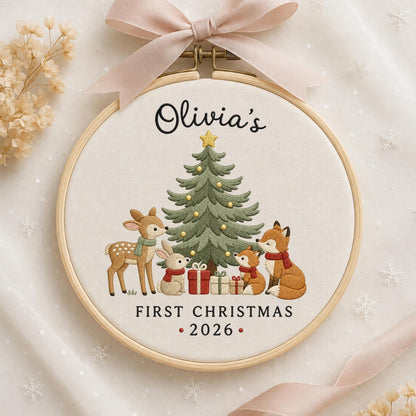 May include: A round wooden embroidery hoop containing fabric with an embroidered green Christmas tree, a deer, a rabbit, and two foxes wearing scarves. The text reads Olivia's FIRST CHRISTMAS 2026. A light pink ribbon is tied at the top of the hoop.