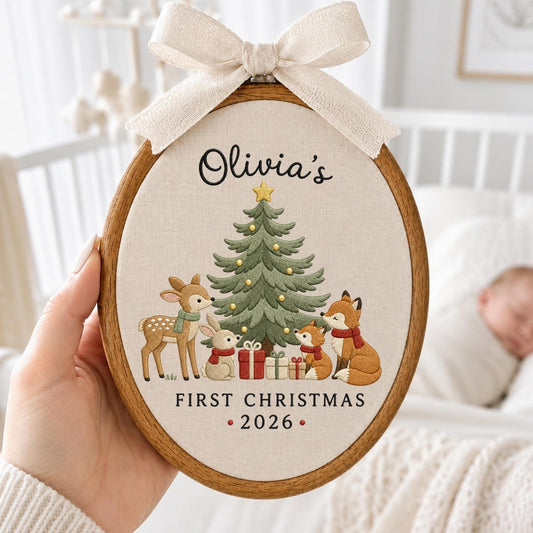 May include: An oval wooden embroidery hoop featuring a cream fabric background with a Christmas tree, a deer, a rabbit, and two foxes. The text reads Olivia's FIRST CHRISTMAS 2026. A beige fabric bow is attached to the top of the wooden frame.