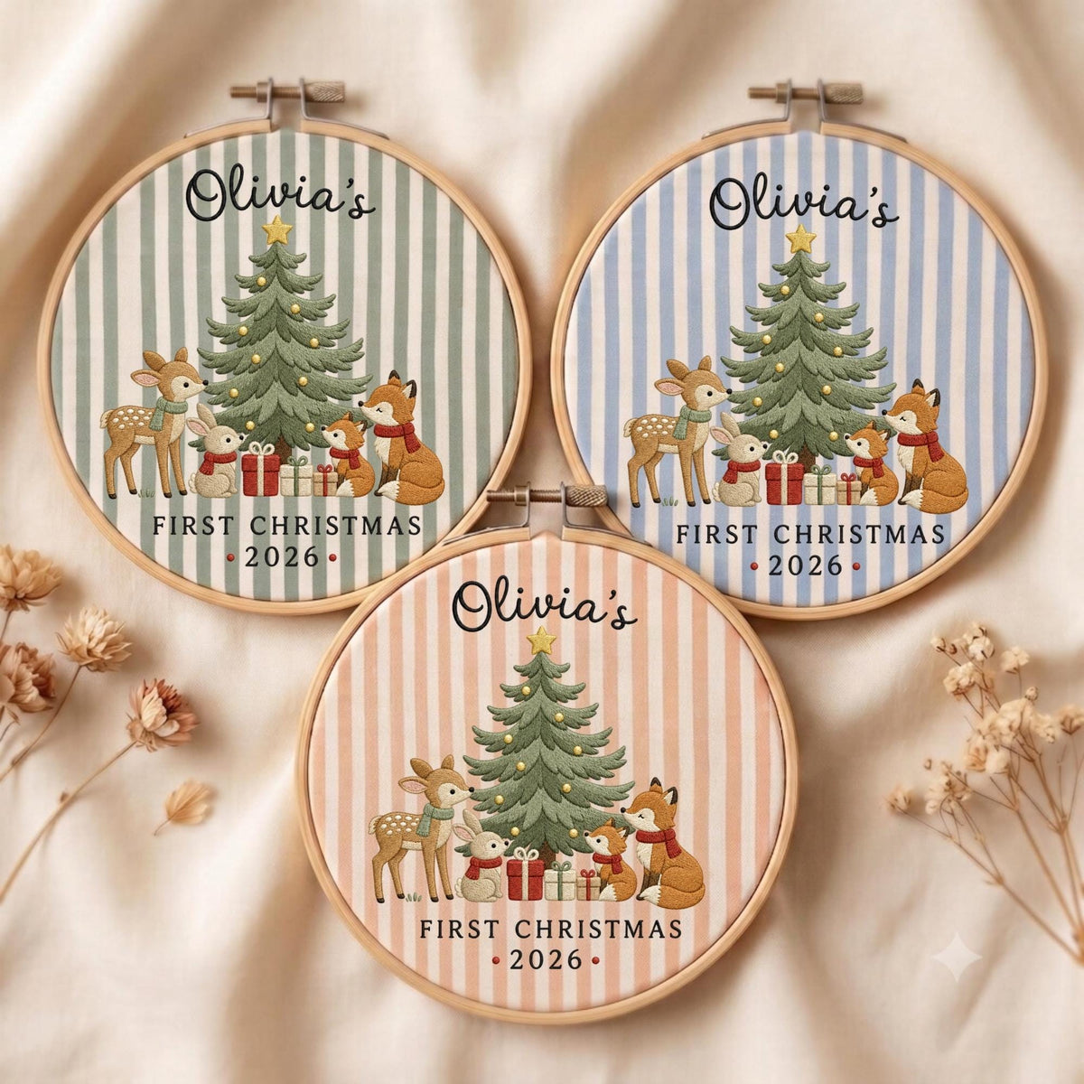 May include: Three circular embroidery hoops featuring striped fabric in sage green, light blue, and peach. Each displays a Christmas tree, a deer, a rabbit, and a fox with gifts. Text reads: Olivia's FIRST CHRISTMAS 2026 in black script and print.