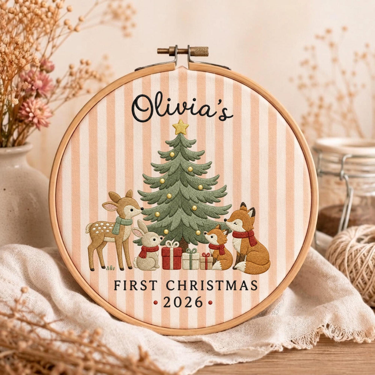May include: A circular wooden embroidery hoop framing fabric with vertical light pink stripes. The design features a green Christmas tree, a deer, a rabbit, and a fox with gifts. Text reads: Olivia's FIRST CHRISTMAS 2026.