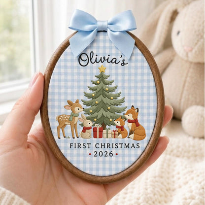 May include: An oval wooden embroidery hoop ornament featuring a light blue and white gingham fabric background. The design includes a green Christmas tree, a deer, a rabbit, and two foxes with the text Olivia's FIRST CHRISTMAS 2026 and a blue bow.
