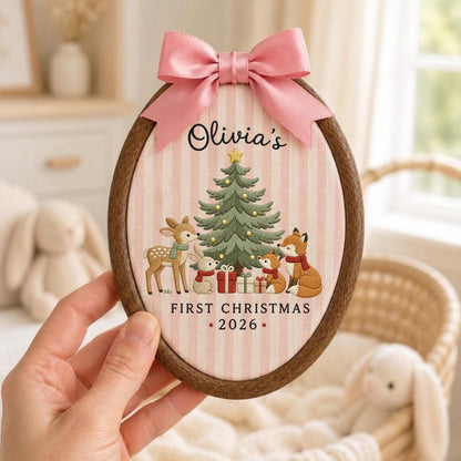 May include: An oval wooden embroidery hoop containing fabric with a pink and white striped pattern. The fabric features an embroidered Christmas tree, a deer, a rabbit, and two foxes with gifts. Text reads: Olivia's FIRST CHRISTMAS 2026. A pink satin bow is attached to the top.