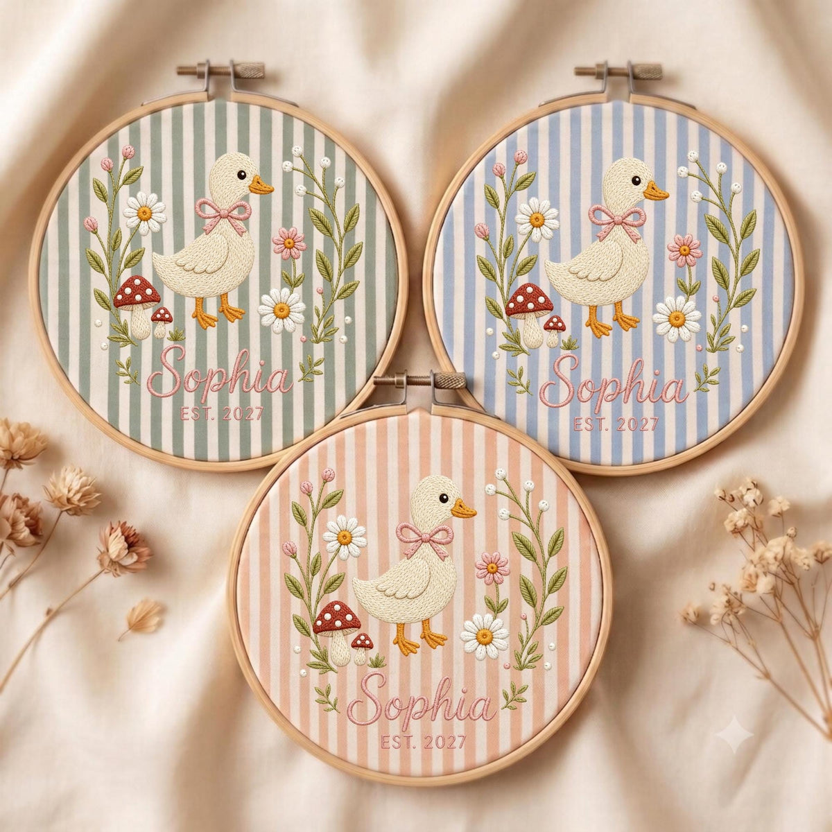 May include: Three circular wooden embroidery hoops featuring fabric with vertical stripes in sage green, light blue, and peach. Each hoop displays an embroidered duck with a pink bow, daisies, mushrooms, and the text Sophia EST. 2027 in pink script.