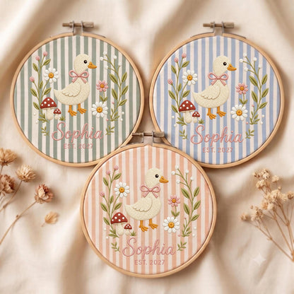 May include: Three circular wooden embroidery hoops featuring fabric with vertical stripes in sage green, light blue, and peach. Each hoop displays an embroidered duck with a pink bow, daisies, mushrooms, and the text Sophia EST. 2027 in pink script.