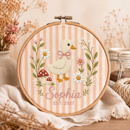 May include: A round wooden embroidery hoop containing fabric with vertical peach and white stripes. The embroidery features a central cream-colored duck with a pink bow, surrounded by white daisies, pink flowers, and red mushrooms with the text Sophia EST. 2027.