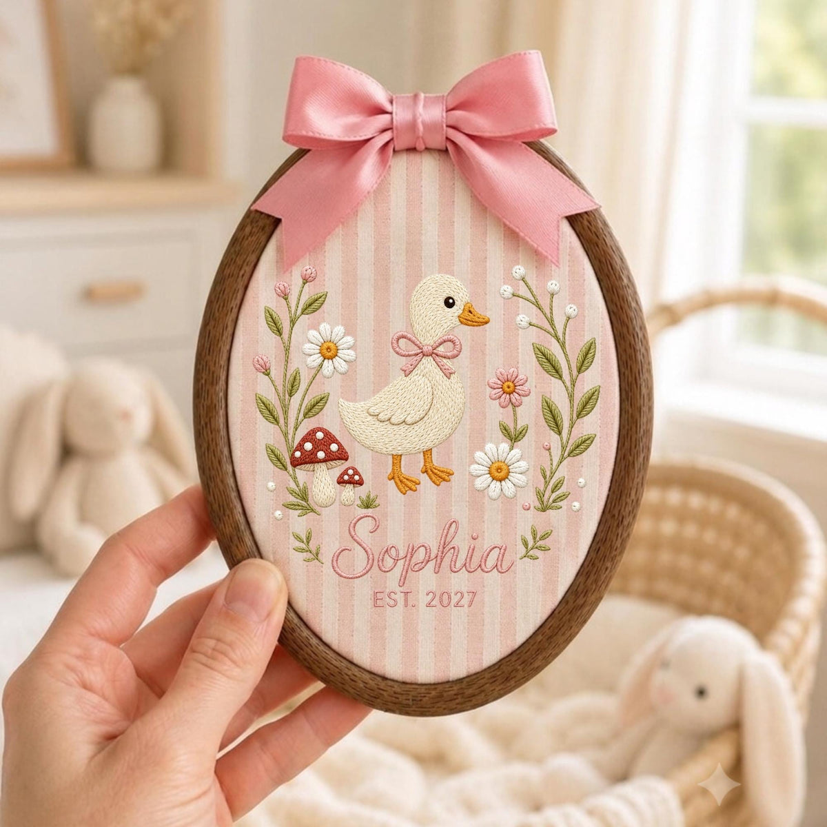 May include: An oval embroidery hoop featuring a light pink and white striped fabric background. The embroidery depicts a cream-colored duck, red mushrooms, and white flowers. The name Sophia and the text EST. 2027 are stitched below. A pink satin bow is attached to the top.
