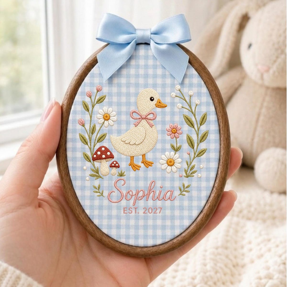 May include: An oval embroidery hoop with a dark wood frame featuring a light blue and white gingham fabric background. The design includes a central embroidered duck, flowers, mushrooms, and the text "Sophia" and "EST. 2027" with a light blue bow.