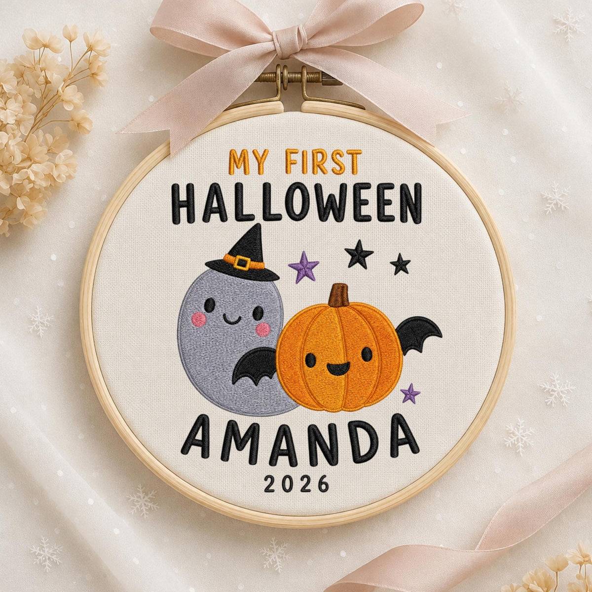 May include: A round wooden embroidery hoop containing white fabric with embroidered text MY FIRST HALLOWEEN AMANDA 2026. The design features a grey ghost wearing a black witch hat and an orange pumpkin with black bat wings and small stars.