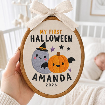 May include: An oval wooden embroidery hoop containing fabric with the embroidered text MY FIRST HALLOWEEN AMANDA 2026. The design features a grey ghost wearing a witch hat and an orange pumpkin with bat wings, accented by small purple and black stars.