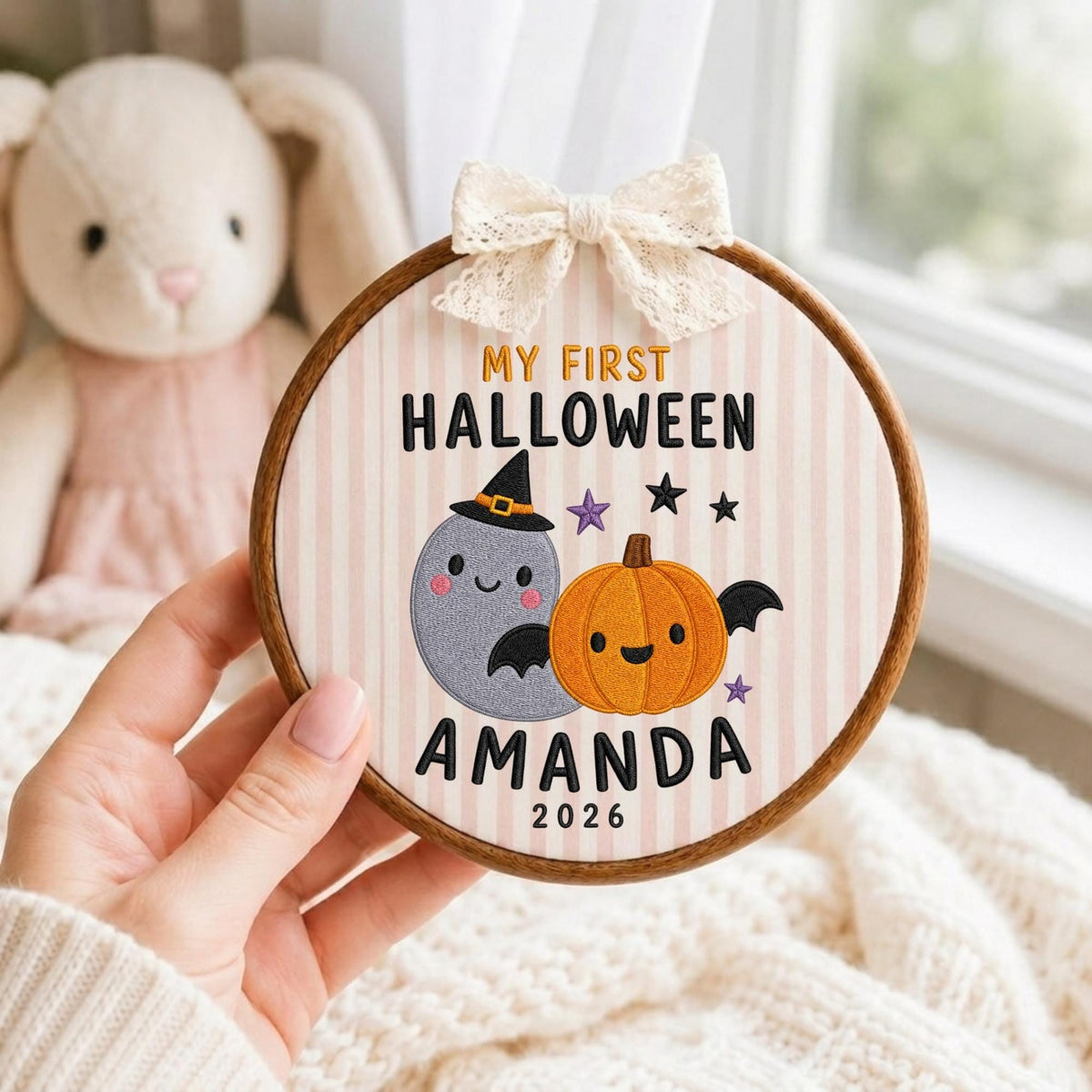 May include: A round wooden embroidery hoop containing fabric with vertical light pink stripes. The embroidery features a gray ghost wearing a witch hat, an orange pumpkin with black bat wings, purple stars, and the text MY FIRST HALLOWEEN AMANDA 2026.