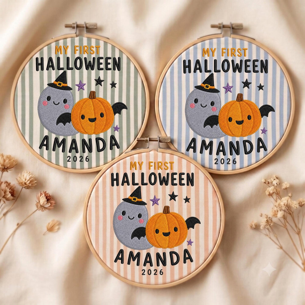 May include: Three circular embroidery hoops featuring striped fabric in sage green, light blue, and peach. Each displays embroidered text MY FIRST HALLOWEEN AMANDA 2026, a smiling grey ghost wearing a black witch hat, an orange pumpkin, and small stars.