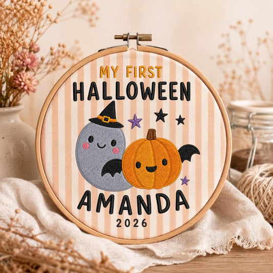 May include: A round wooden embroidery hoop containing fabric with vertical beige stripes. The embroidery features a gray ghost wearing a black witch hat, an orange pumpkin with black wings, purple stars, and the text MY FIRST HALLOWEEN AMANDA 2026.