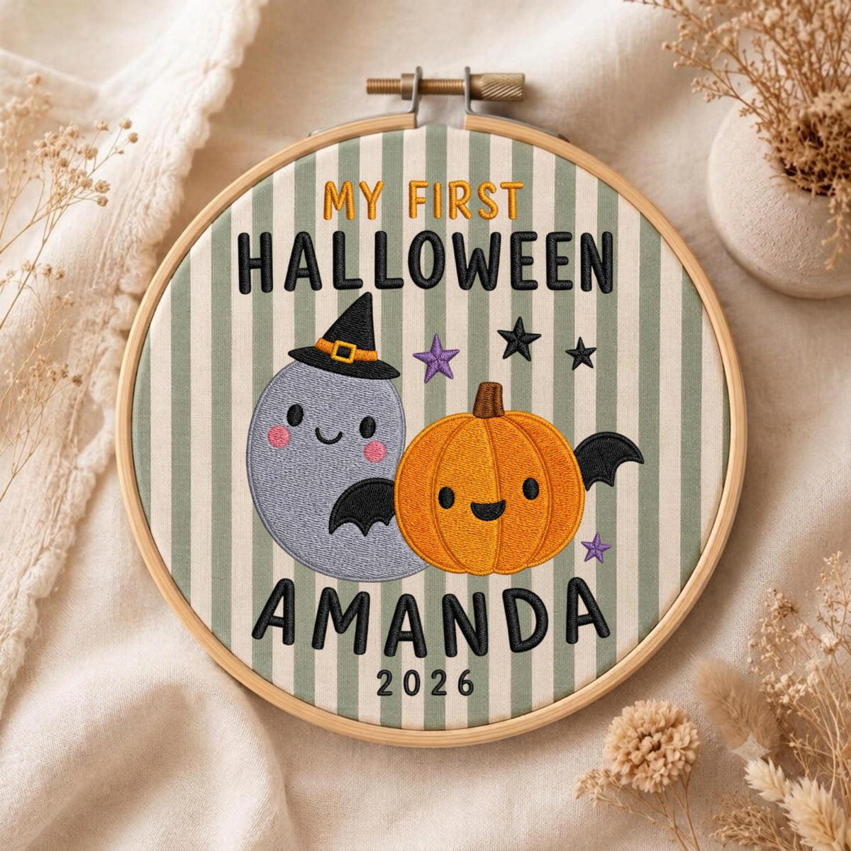 May include: A round wooden embroidery hoop containing fabric with green and white vertical stripes. The embroidery features a grey ghost wearing a black witch hat, an orange pumpkin with black wings, purple and black stars, and the text MY FIRST HALLOWEEN AMANDA 2026.