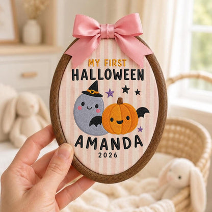 May include: An oval wooden embroidery hoop containing fabric with vertical pink stripes. It features embroidered text reading MY FIRST HALLOWEEN AMANDA 2026, a gray ghost wearing a witch hat, an orange pumpkin with bat wings, and small stars. A pink satin bow is attached to the top.