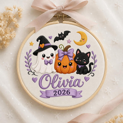 May include: A round wooden embroidery hoop containing a white fabric panel with a stitched design of a ghost in a witch hat, a pumpkin with a purple bow, and a black cat. The text Olivia and 2026 are embroidered in purple. A light pink ribbon is tied at the top.