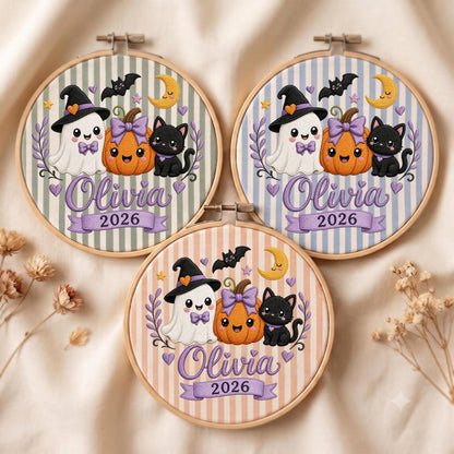 May include: Three circular embroidery hoops featuring a Halloween design with a ghost in a witch hat, a pumpkin with a purple bow, and a black cat. Each hoop displays the name Olivia and the year 2026 in purple script on a striped fabric background.