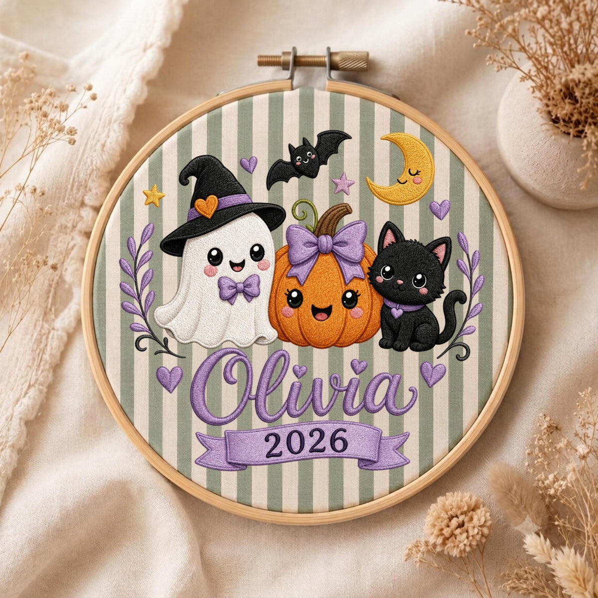 May include: A round wooden embroidery hoop containing fabric with green and white vertical stripes. The design features a white ghost in a black hat, an orange pumpkin with a purple bow, and a black cat. Text reads Olivia and the year 2026.