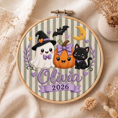 May include: A round wooden embroidery hoop containing fabric with green and white vertical stripes. The design features a white ghost in a black hat, an orange pumpkin with a purple bow, and a black cat. Text reads Olivia and the year 2026.